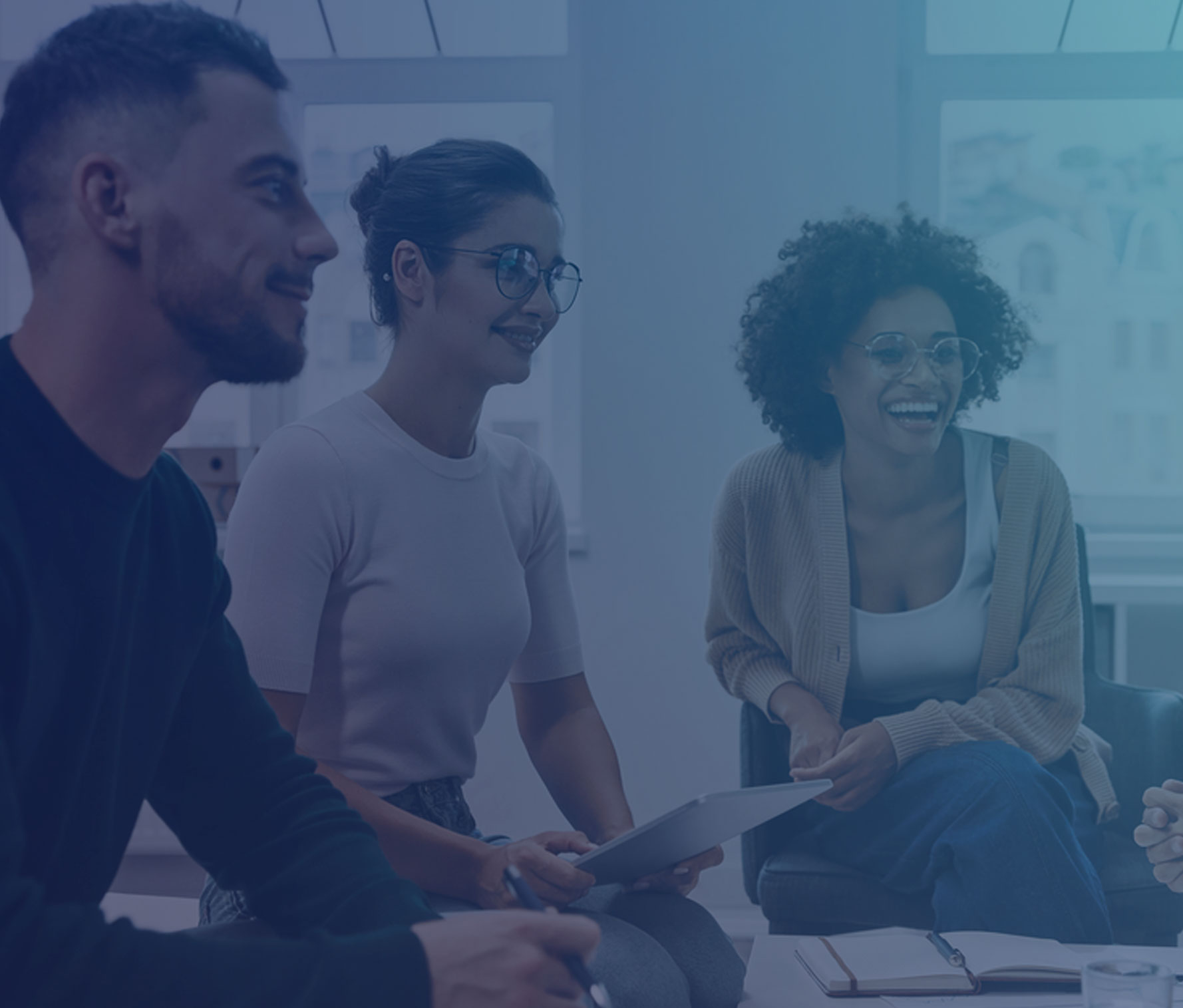 A diverse group of young professionals in a bright office space having a casual meeting. They are sitting in a circle and appear to be engaged in a lively discussion. One person holds a tablet while others are smiling and participating in the conversation. The office has large windows and natural lighting.